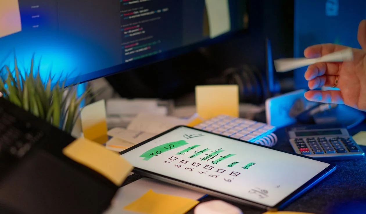 A software workspace with code on a monitor, a planning tablet and desk supplies.
