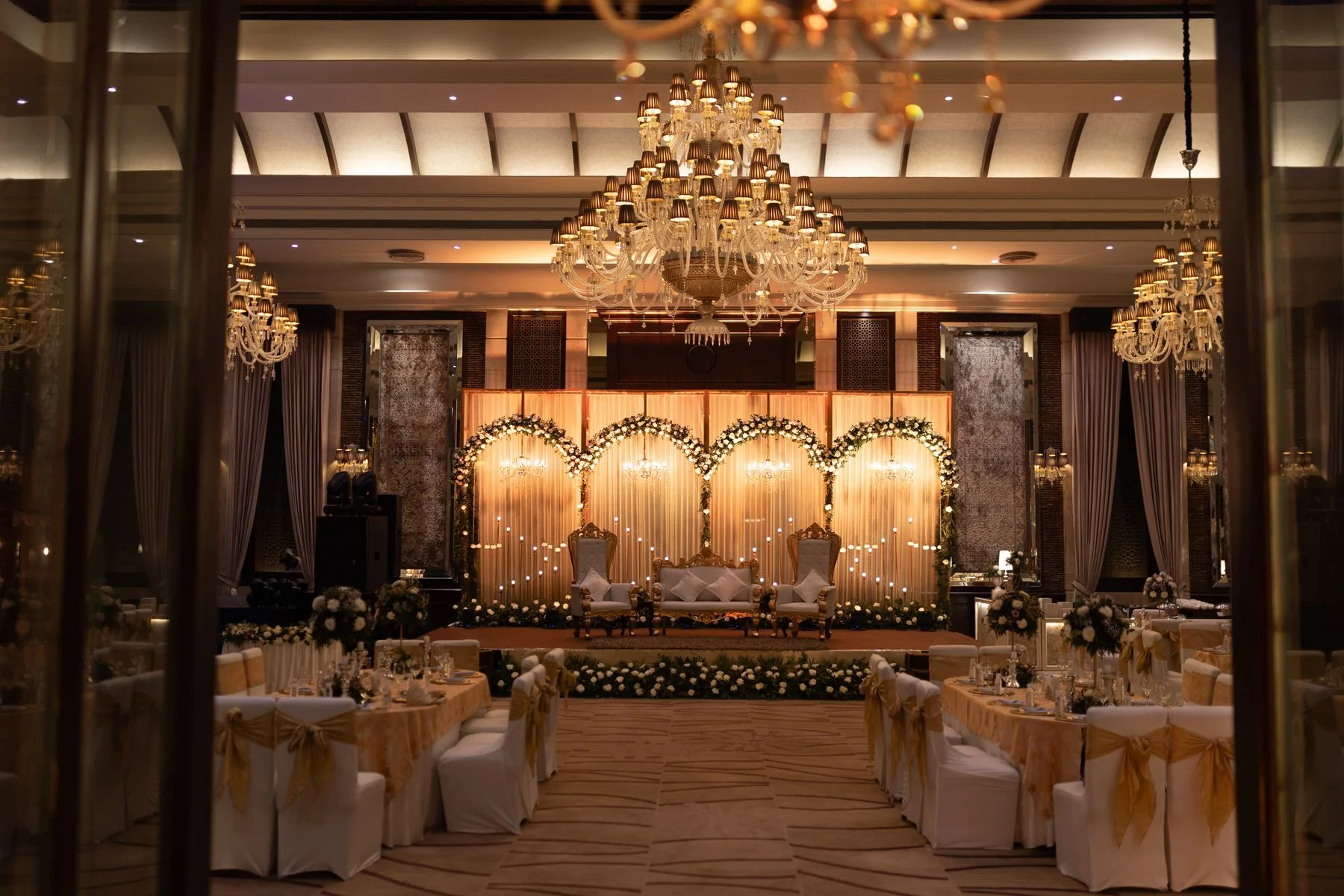 An elegant reception hall with crystal chandeliers, white banquet seating and an illuminated floral stage.