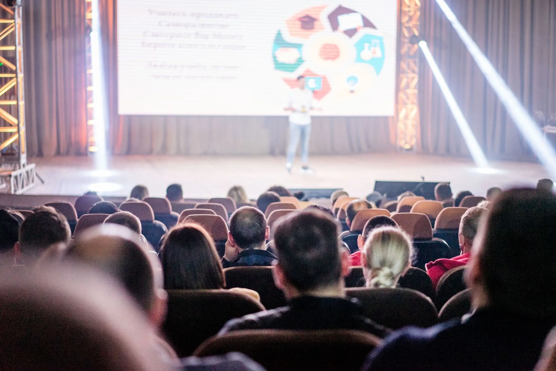 Audience members seated in an auditorium face a speaker on a brightly lit conference stage.