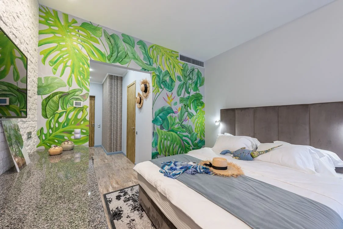 A hotel bedroom with white bedding, a straw hat and a green tropical leaf mural.