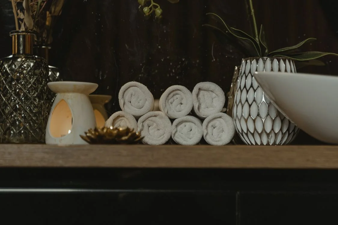 Rolled white towels, ceramic vessels and an oil warmer on a dark wooden spa shelf.